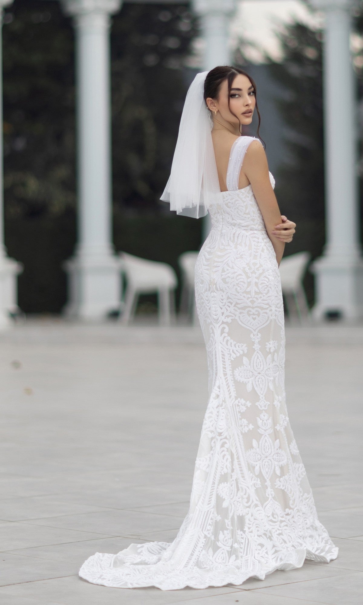 A woman in the Off-Shoulder White Bridal Gown: Tina Holly TW103 stands outdoors, looking over her shoulder, with white columns and blurred greenery in the background.